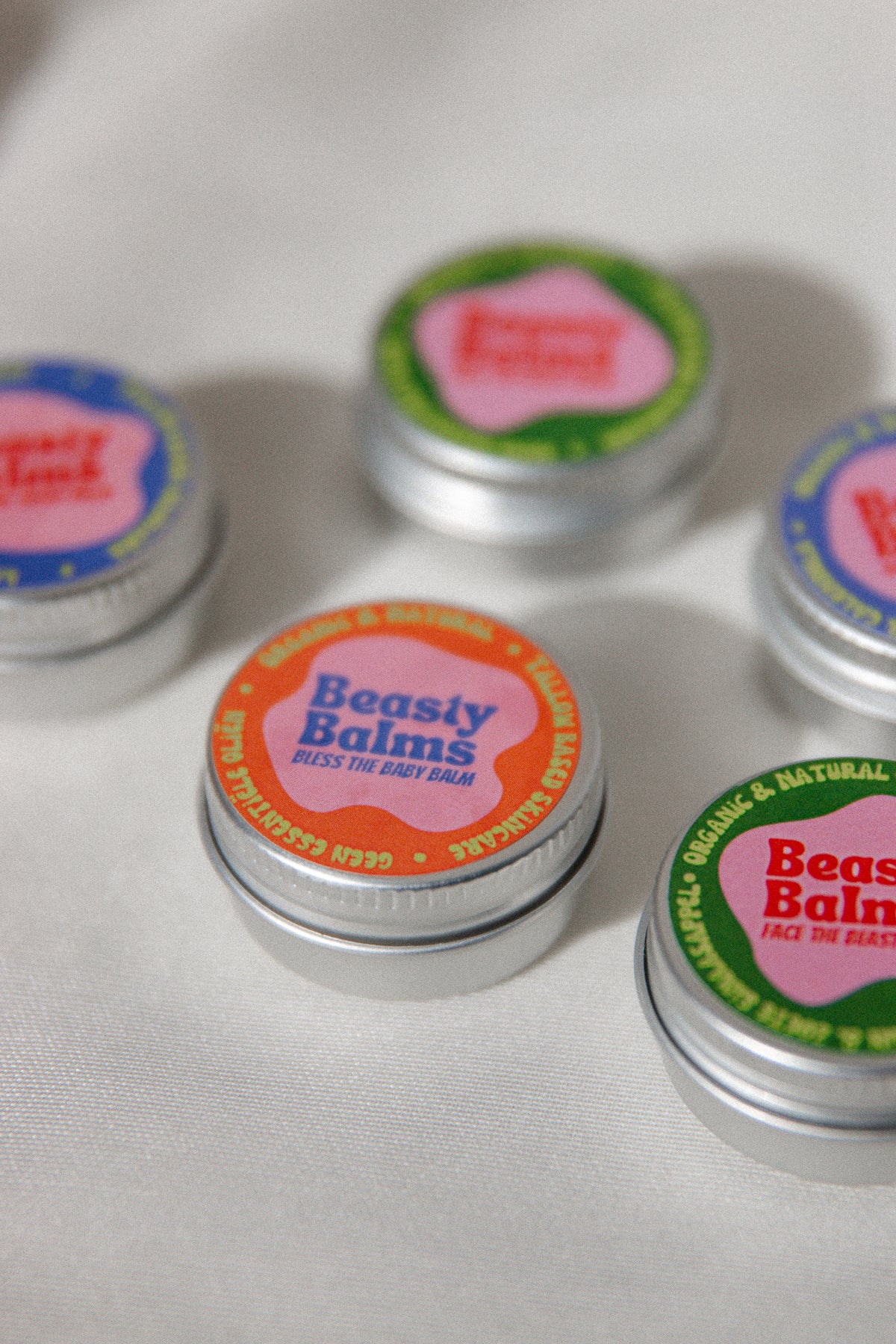 Close-up of the Bless the Baby mini tin with its orange and pink label, other Beasty Balms tins softly blurred in the background.