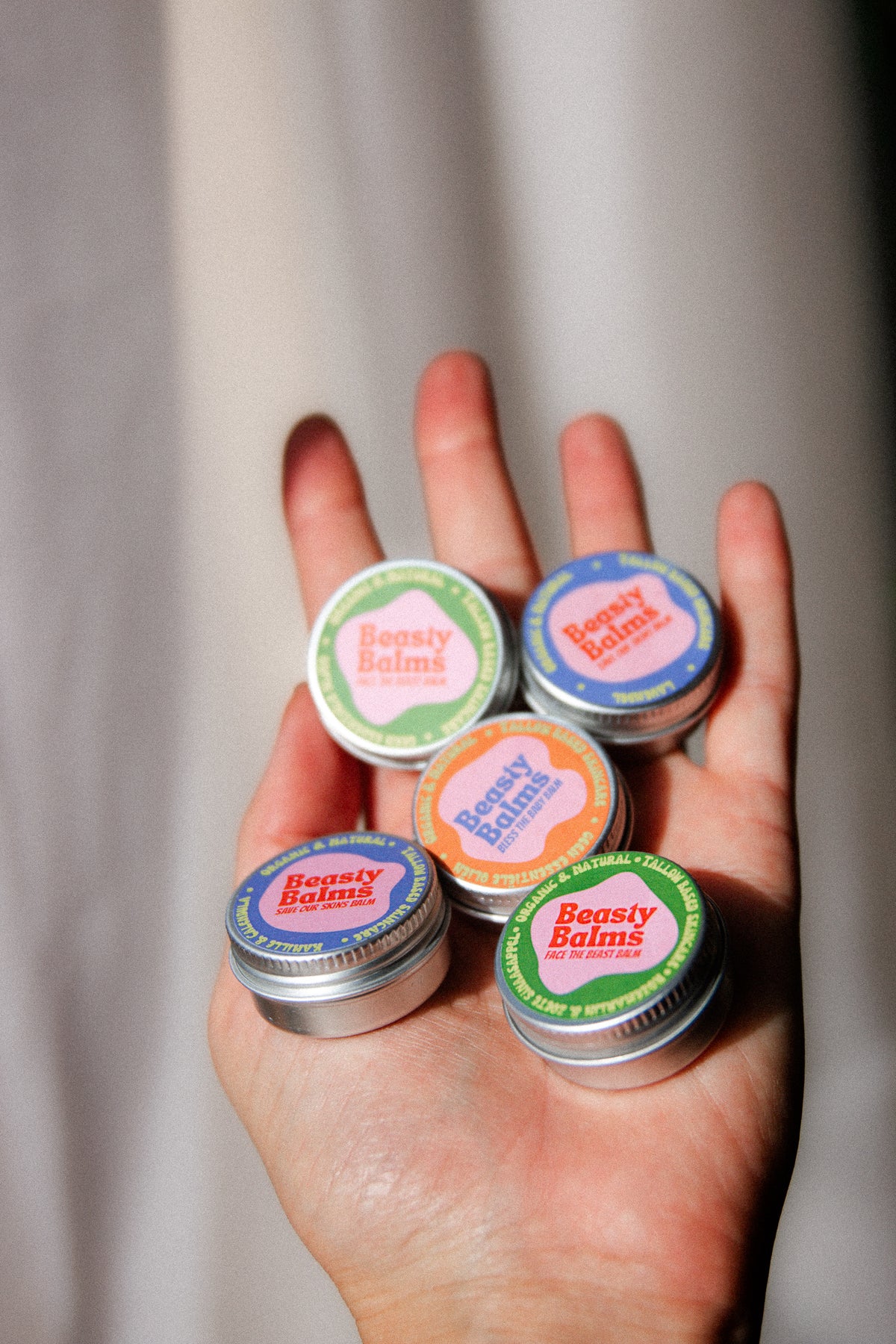 A hand holding five colourful Beasty Balms mini tins, the full tallow balm sample set.